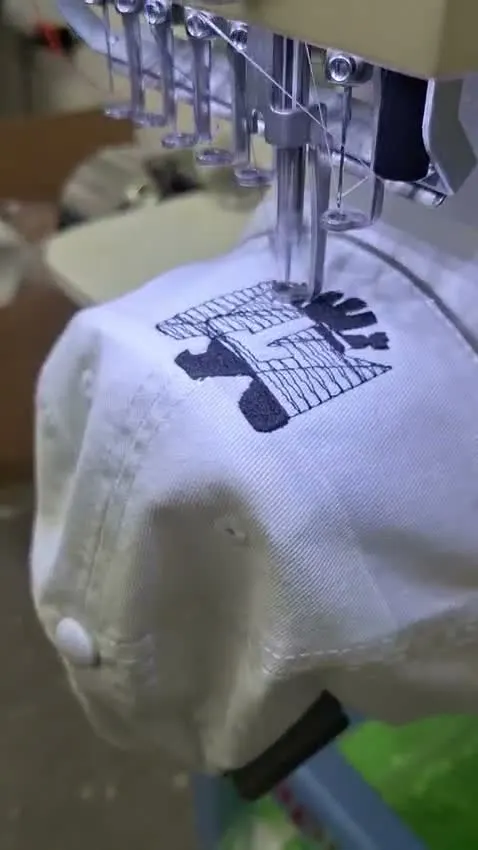 Máquina bordando un logo en hilo negro sobre una gorra blanca